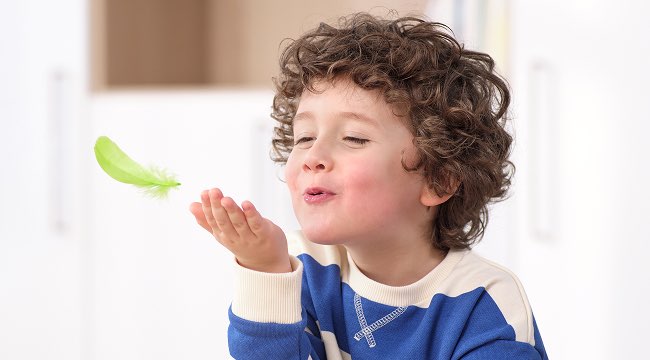 Kind pustet eine grüne Feder in die Luft bei spielerischer Atemübung